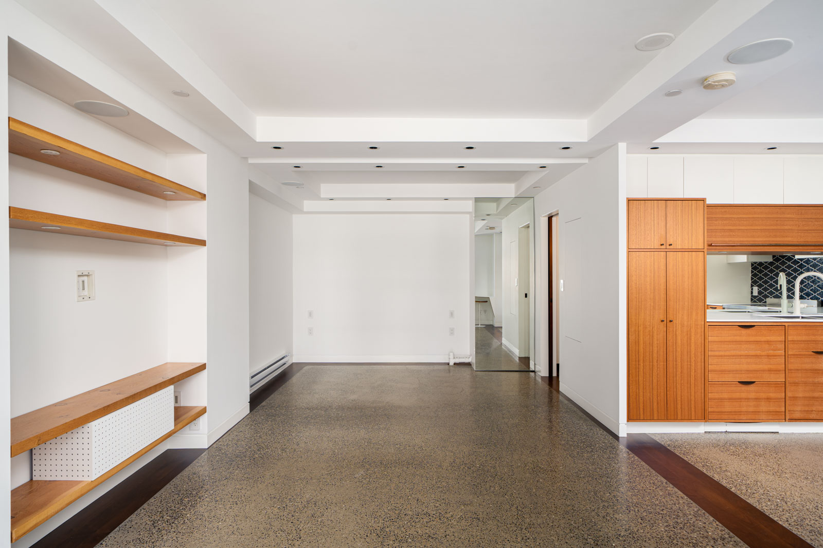 Modern, unfurnished apartment interior with white walls, open shelves, polished concrete floor, and a wooden kitchen area visible on the right side.