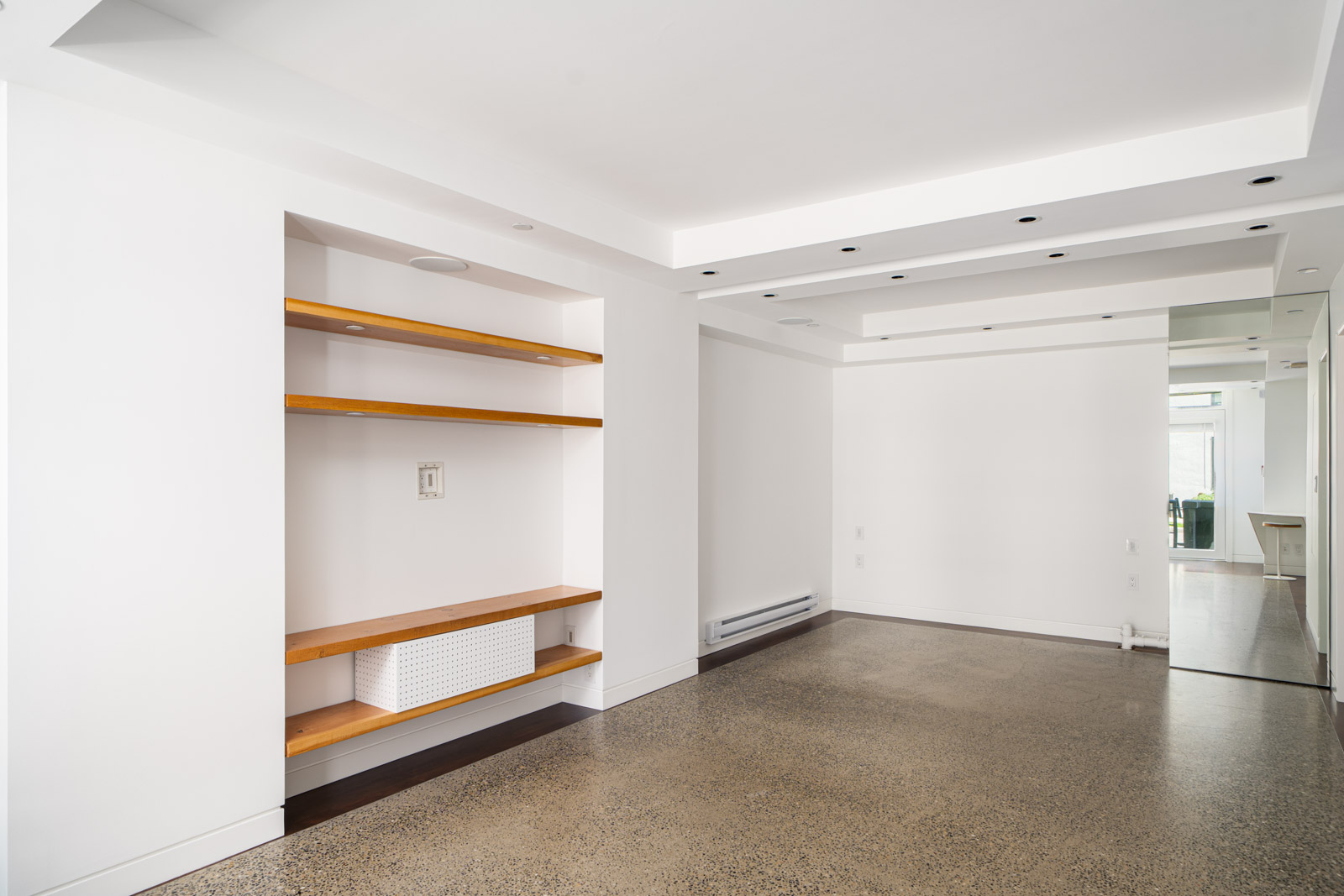 Empty modern living room with white walls, built-in wooden shelves, recessed lighting, and polished concrete floor leading to a hallway and kitchen area in the background.