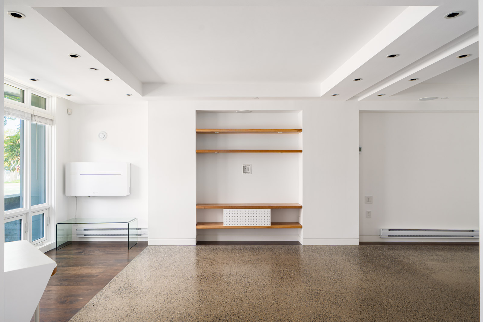 A modern, empty room with white walls, built-in wooden shelves, large windows, and polished concrete floors.
