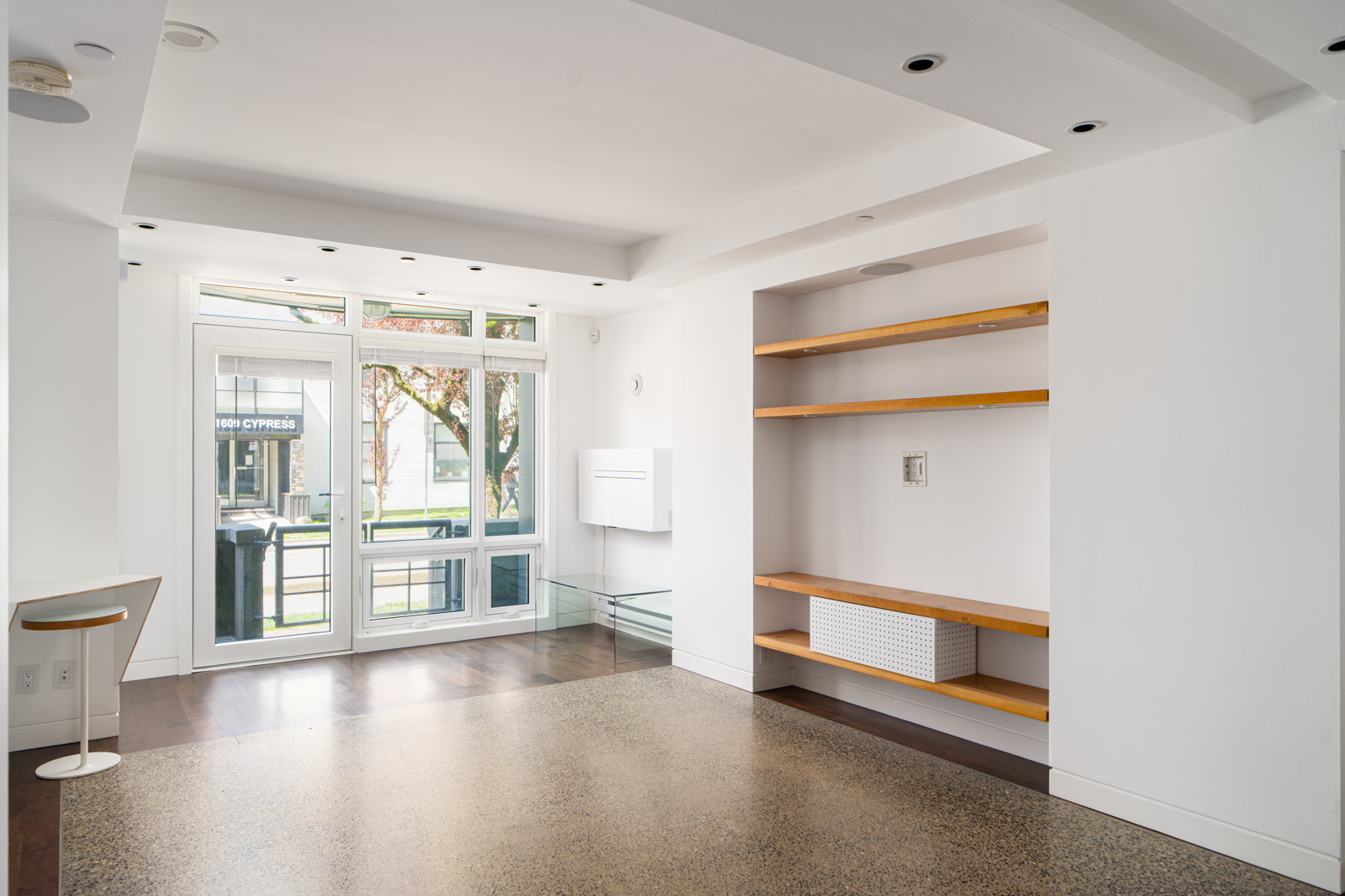 A modern, empty living room with white walls, built-in wooden shelves, large window, glass door, and polished concrete floors.
