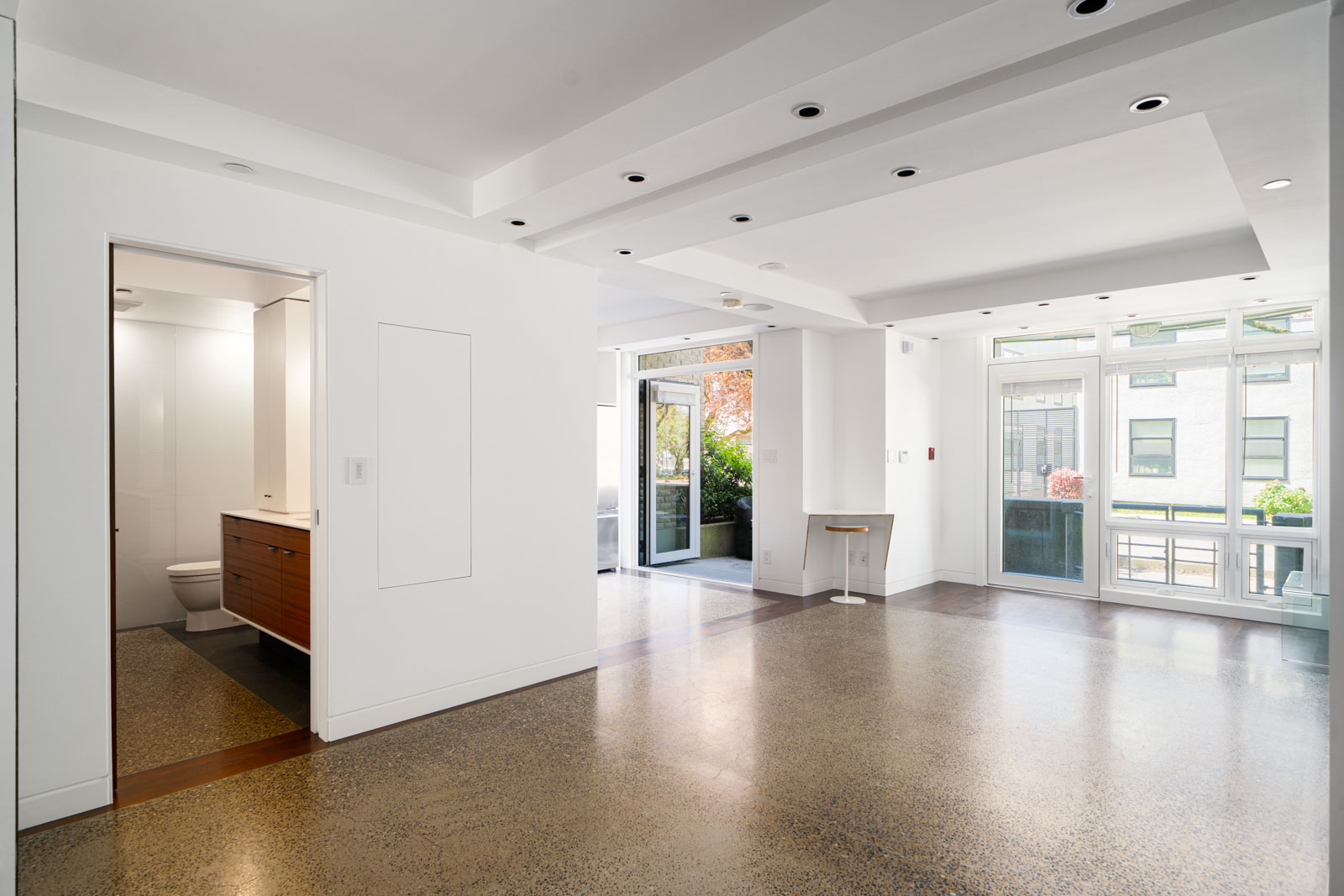 A modern, unfurnished open-plan living space with large windows, polished concrete floors, recessed lighting, and a visible bathroom to the left.