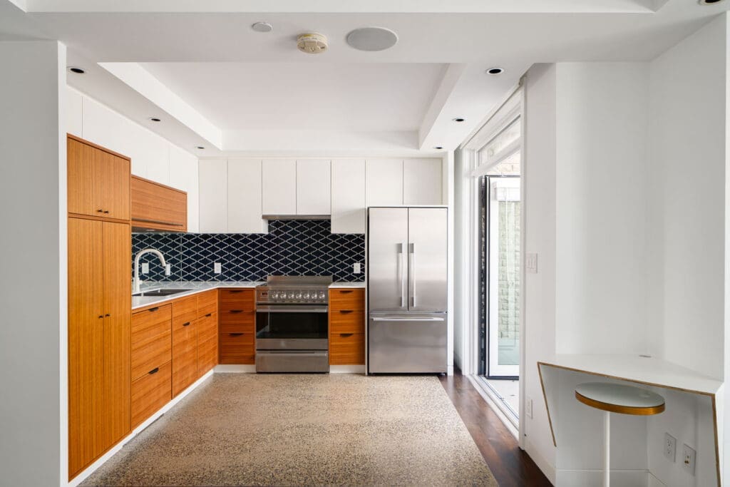 Modern kitchen with wood and white cabinets, stainless steel appliances, dark geometric tile backsplash, and a built-in desk with a stool near a glass door.
