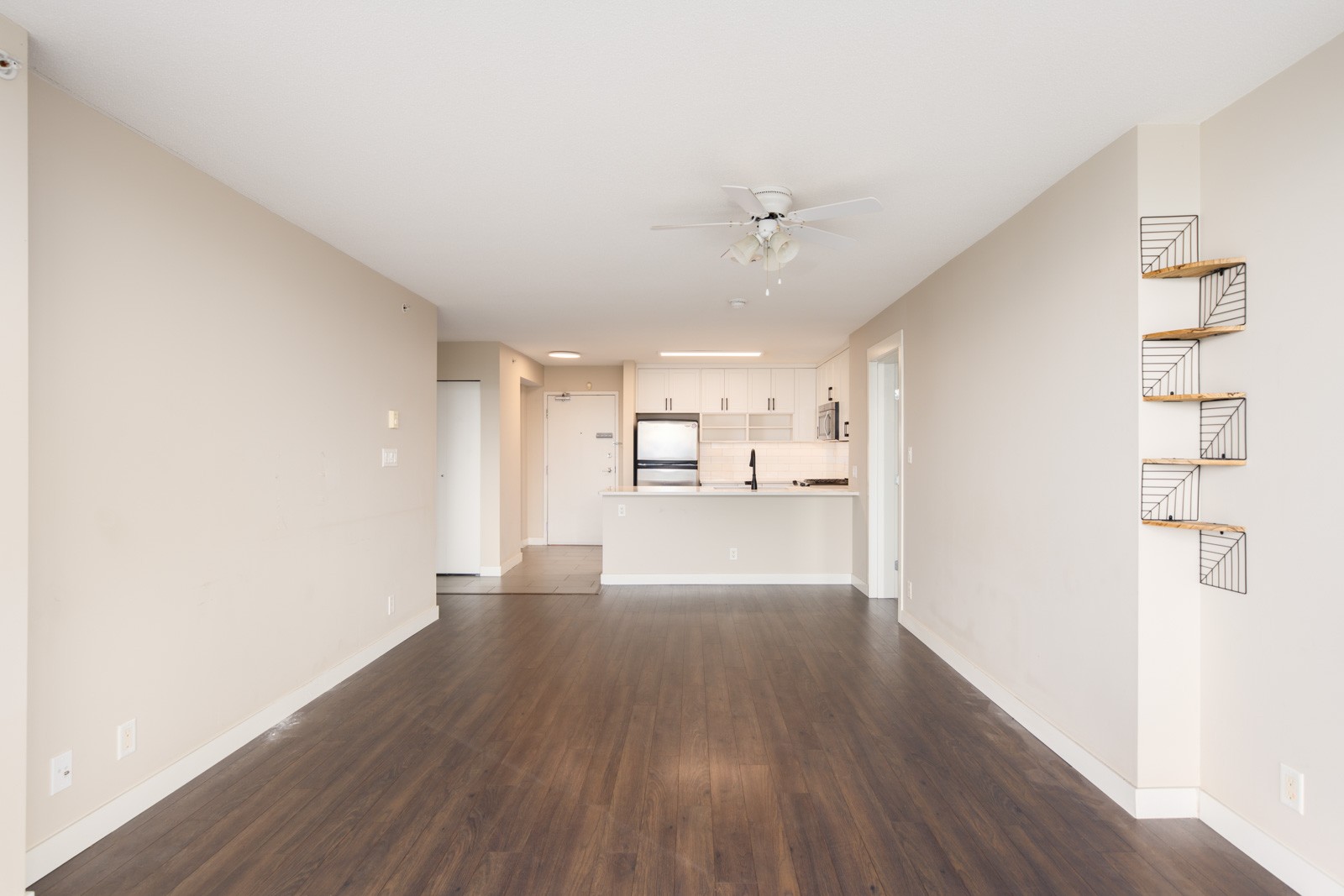 Empty apartment with beige walls, dark wood flooring, a ceiling fan, built-in shelves on the wall, and a view of a white kitchen with stainless steel appliances in the background.