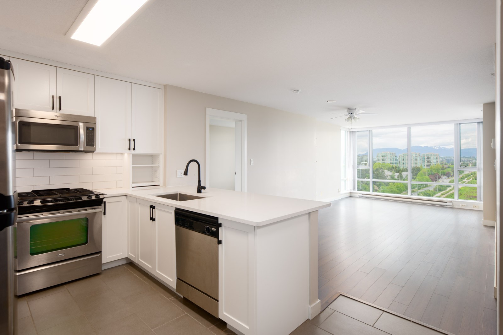 Modern kitchen with stainless steel appliances and white cabinets opens to a bright living area with large windows and a city view.