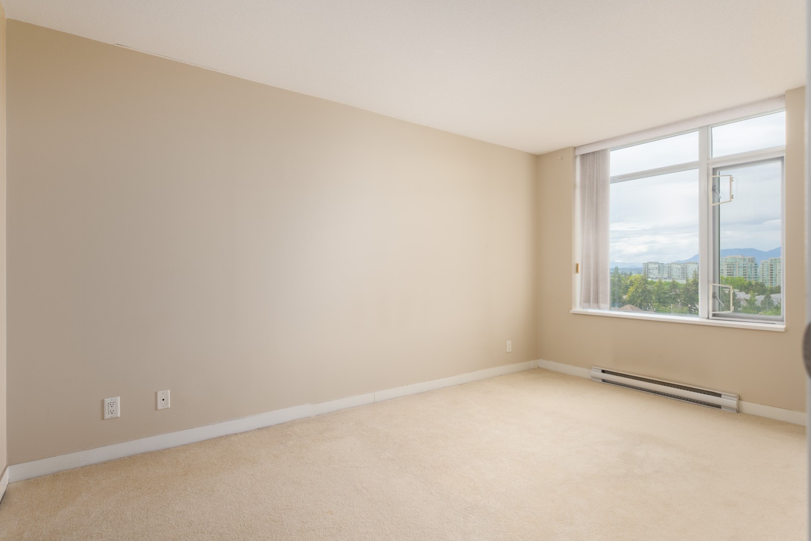 Empty beige-walled room with carpeted floor, large window, baseboard heater, and cityscape visible outside. Two electrical outlets on the left wall.