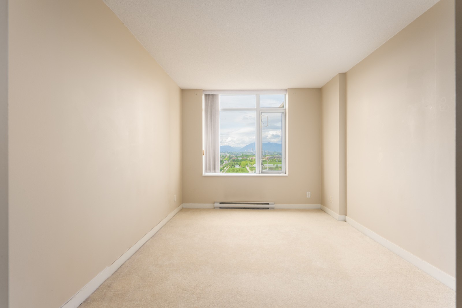 Empty beige room with carpet flooring, cream walls, and a large window at the far end offering a view of greenery and mountains outside.