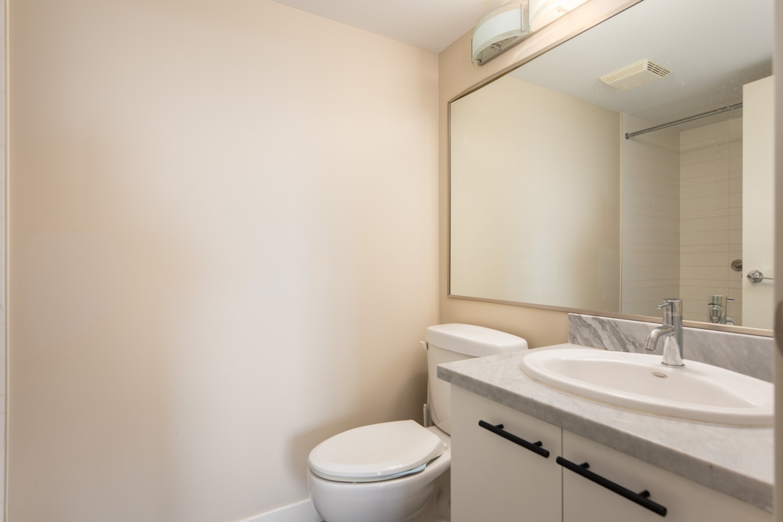 A simple bathroom with a toilet, a sink with a marble countertop, a large wall mirror, and a partial view of a shower area.