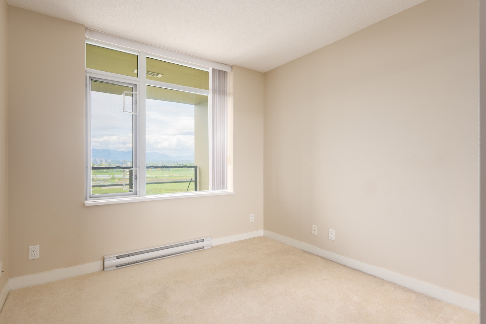 Empty beige room with carpet flooring, a large window, and a view of green fields and mountains outside. White baseboard heater and electrical outlets are visible on the walls.