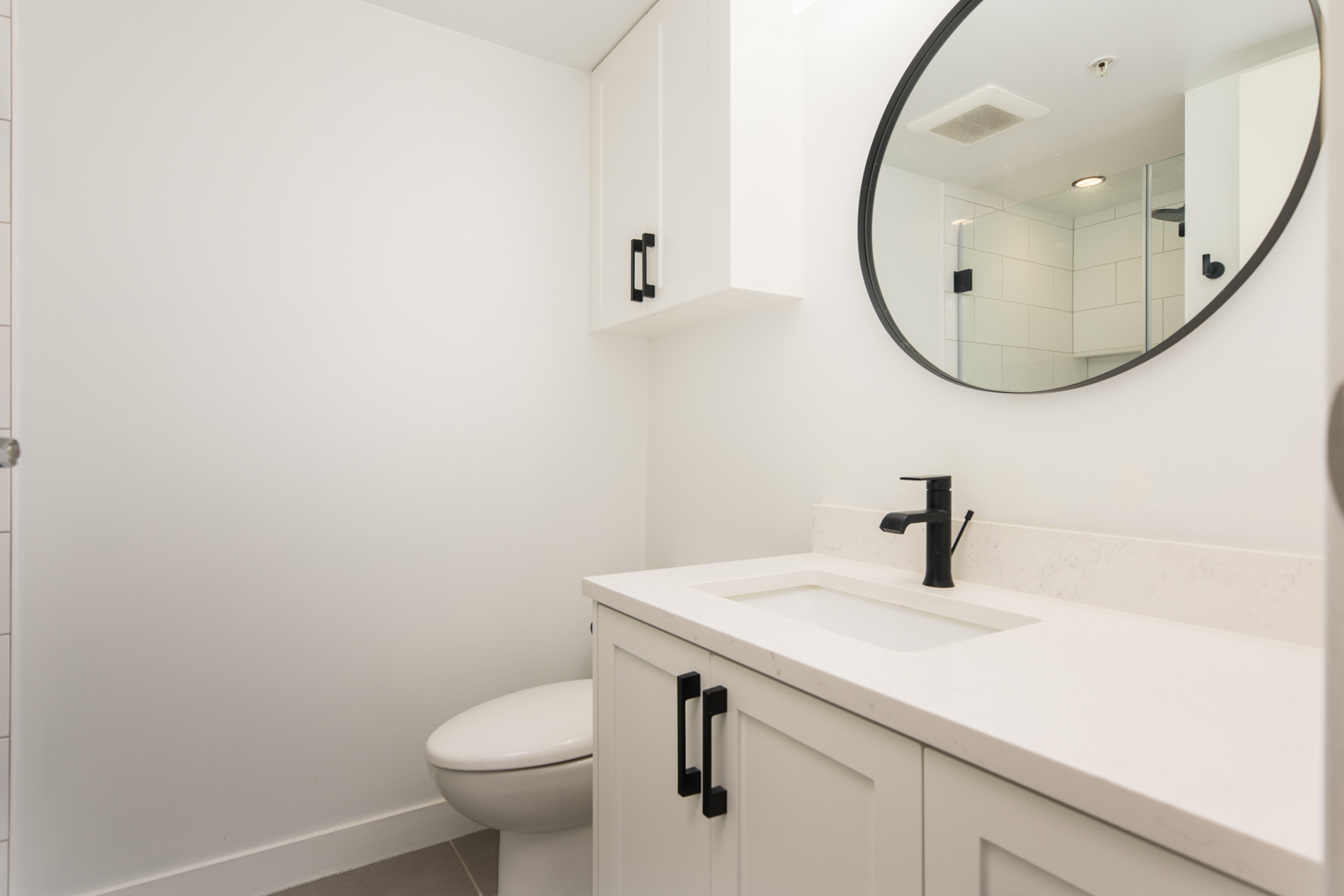 Modern bathroom with white walls, a toilet, a white vanity with black fixtures, a round mirror, and a partial view of a glass shower.