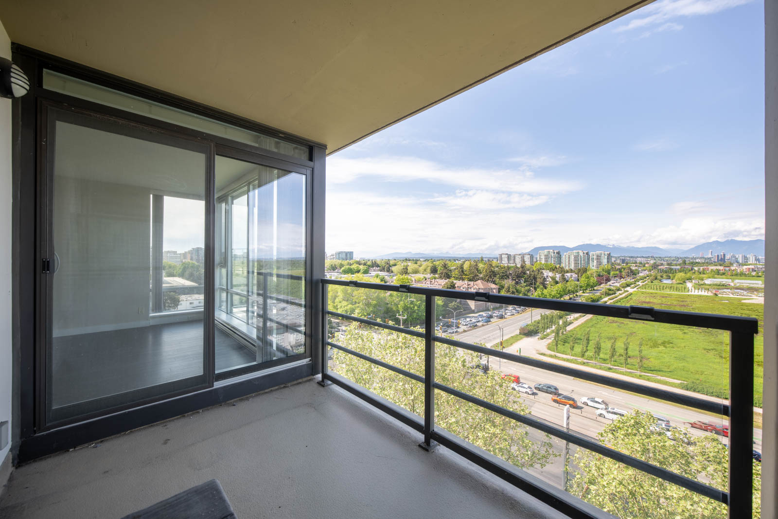A high-rise apartment balcony with glass railing overlooks a cityscape, green park, and distant mountains under a partly cloudy sky.