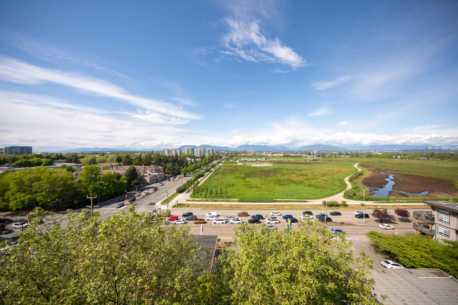 Aerial view of a cityscape with a busy road, parked cars, green fields, trees, and distant buildings under a partly cloudy blue sky.