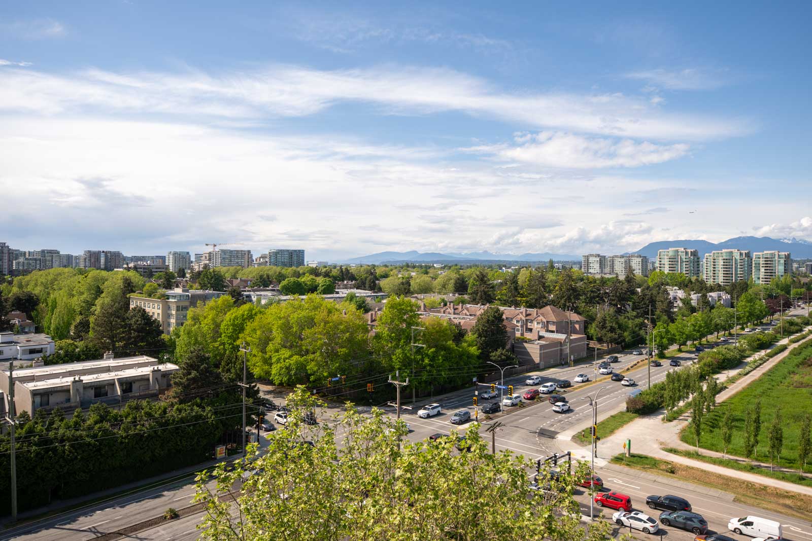 Aerial view of a busy urban intersection surrounded by trees, residential buildings, and distant mid-rise apartments under a partly cloudy sky.