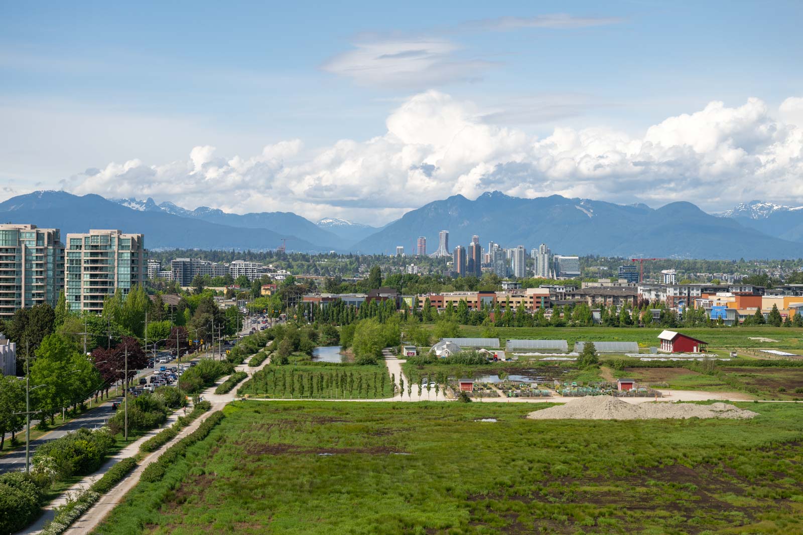 A cityscape with mid-rise buildings, a farm, green fields, and distant mountains under a partly cloudy sky.