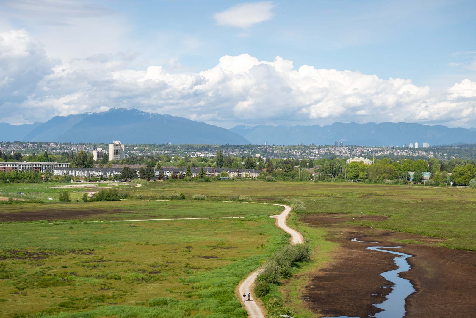 Wide view of a green park with a winding dirt path, small creek, and distant city buildings under a partly cloudy sky with mountains in the background.