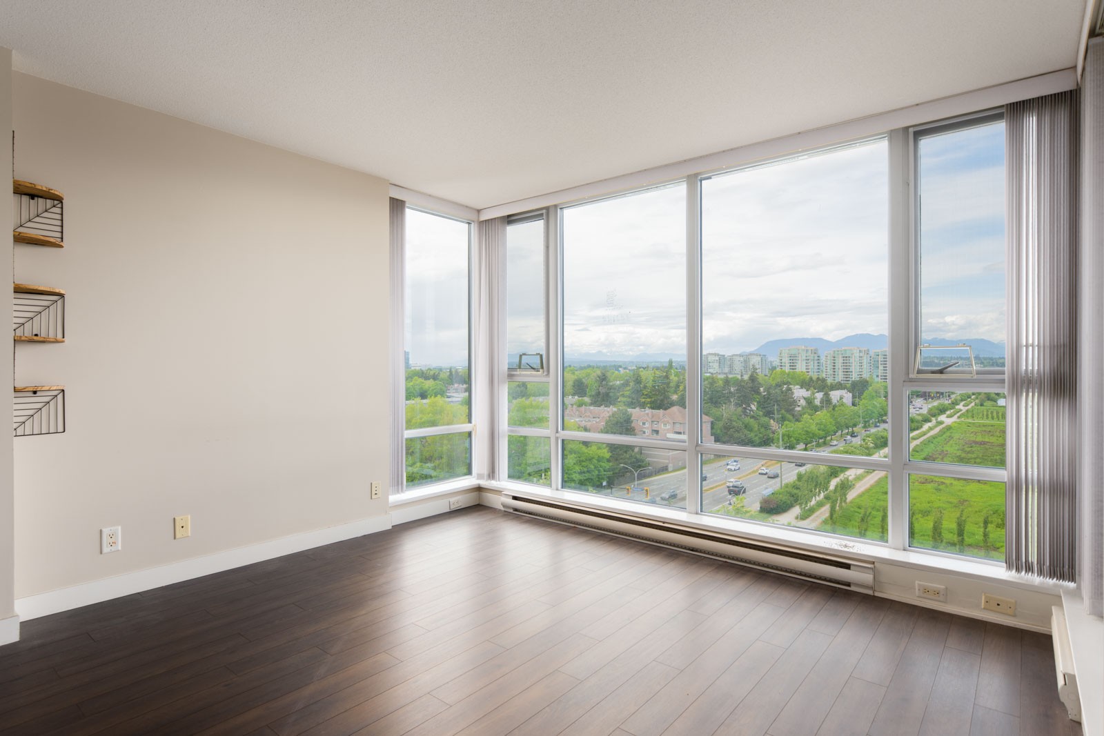 Empty room with large floor-to-ceiling windows, dark wood flooring, neutral walls, and a view of city buildings and greenery outside.