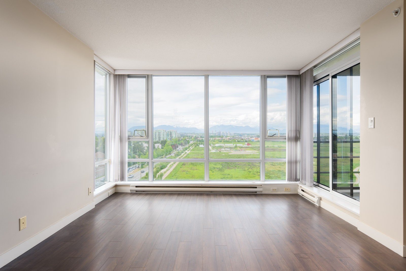 Empty room with hardwood floors, large corner windows, and a view of green fields, city buildings, and mountains under a cloudy sky.