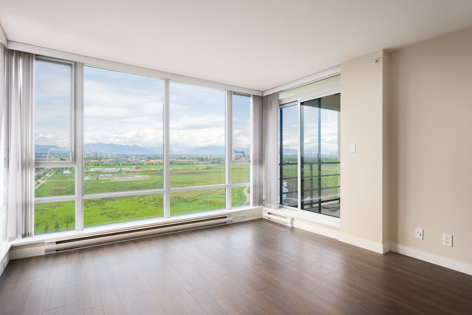 Empty room with large floor-to-ceiling windows, wood flooring, neutral walls, and a sliding glass door leading to a balcony with a view of green fields and distant mountains.