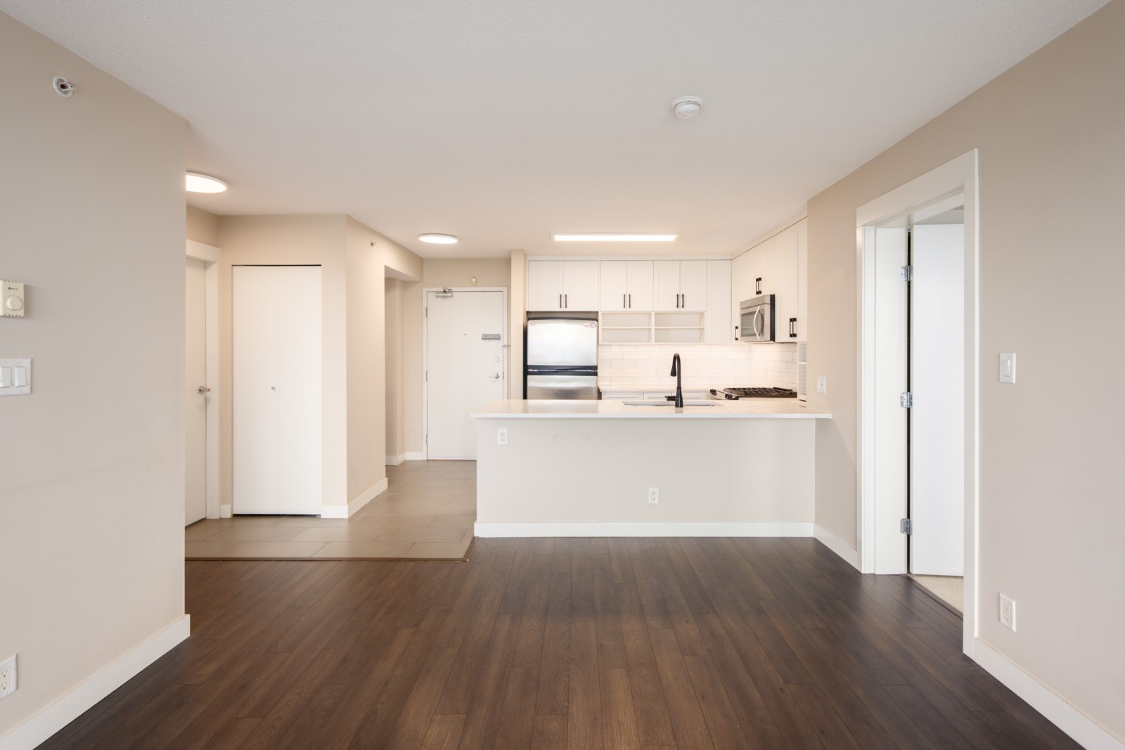 Modern apartment interior with an open kitchen, stainless steel appliances, white cabinets, wood flooring, and neutral-colored walls.