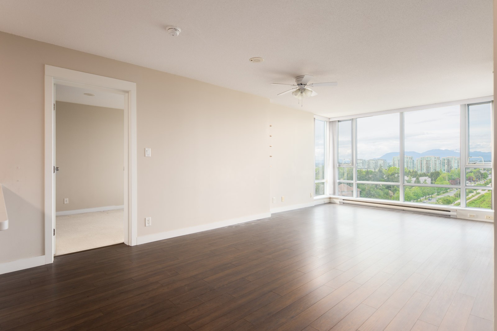 Empty living room with dark wood flooring, beige walls, ceiling fan, large windows, and a view of greenery and buildings outside. A doorway leads to another room.