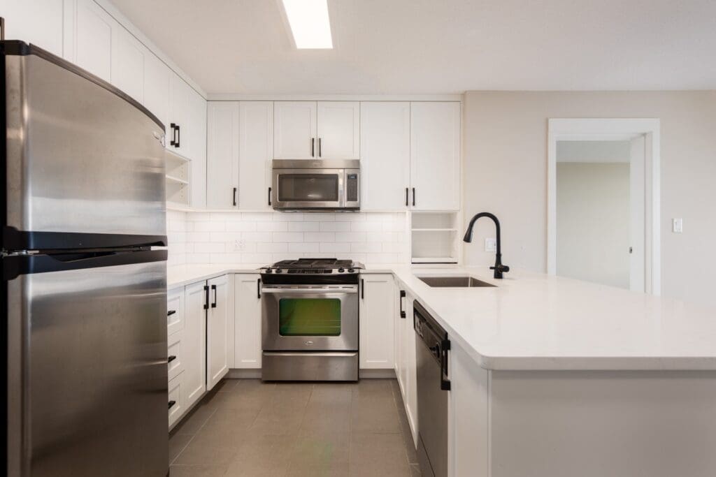 Modern kitchen with white cabinets, stainless steel appliances, black fixtures, and a white countertop under bright overhead lighting.