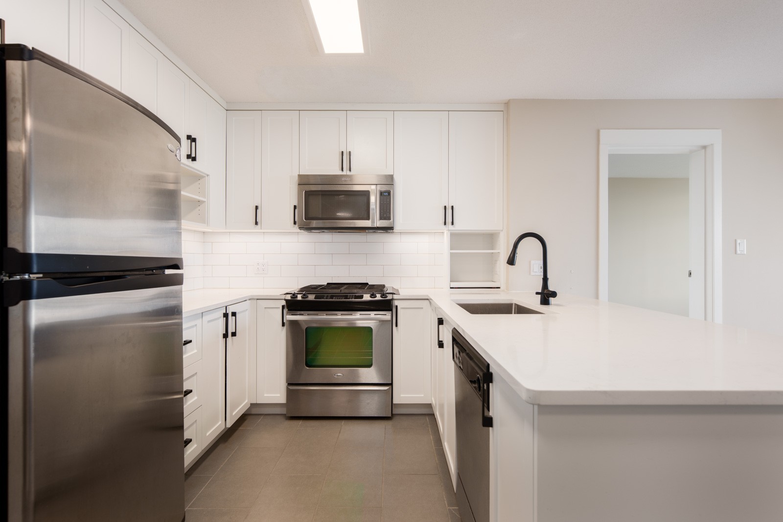 Modern kitchen with white cabinets, stainless steel appliances, black fixtures, and a white countertop under bright overhead lighting.