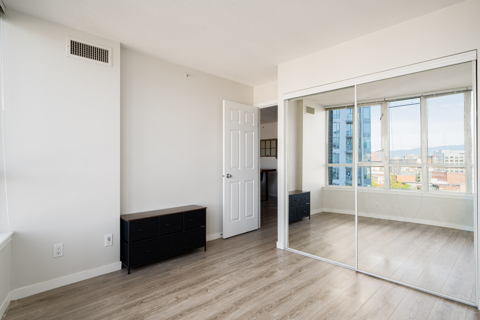 Bright, empty bedroom with light wood flooring, mirrored closet doors, white walls, a small black dresser, and large windows showing a city view.