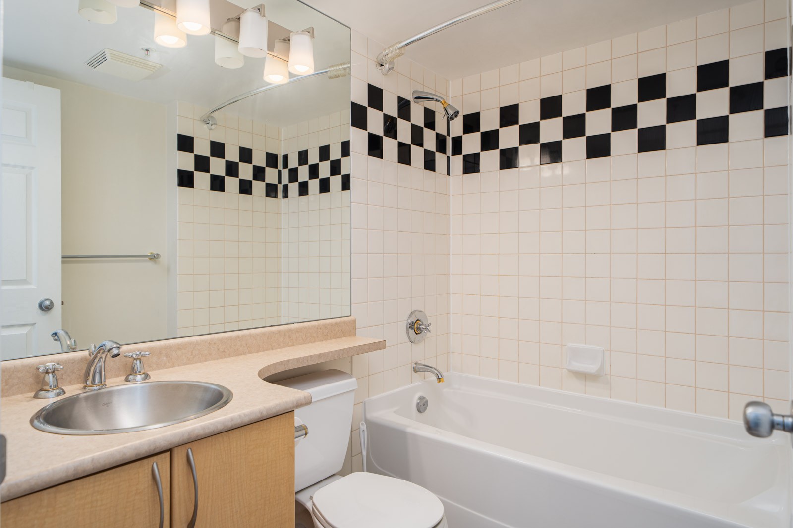 Bathroom with a white bathtub, wall-mounted shower, single sink vanity, large mirror, and white tiles featuring a black checkerboard border.