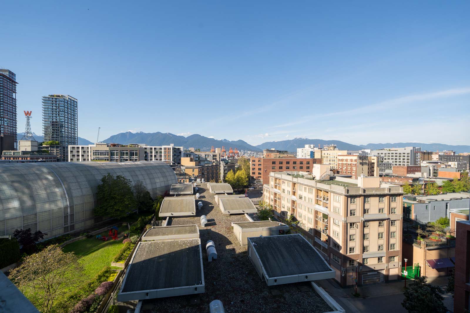 Urban cityscape with mid-rise buildings, a curved structure on the left, and mountains visible in the background under a clear blue sky.