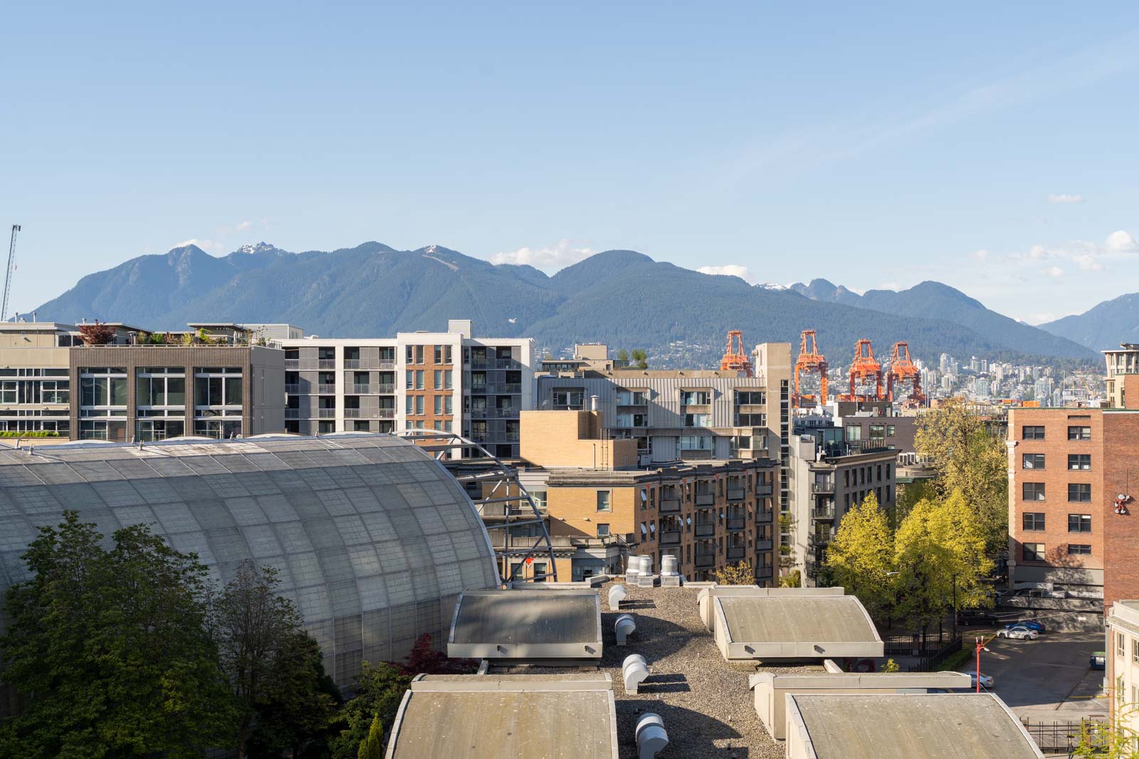 Urban landscape with mid-rise buildings, a curved-roof structure, orange industrial cranes, trees, and mountains in the background under a clear blue sky.