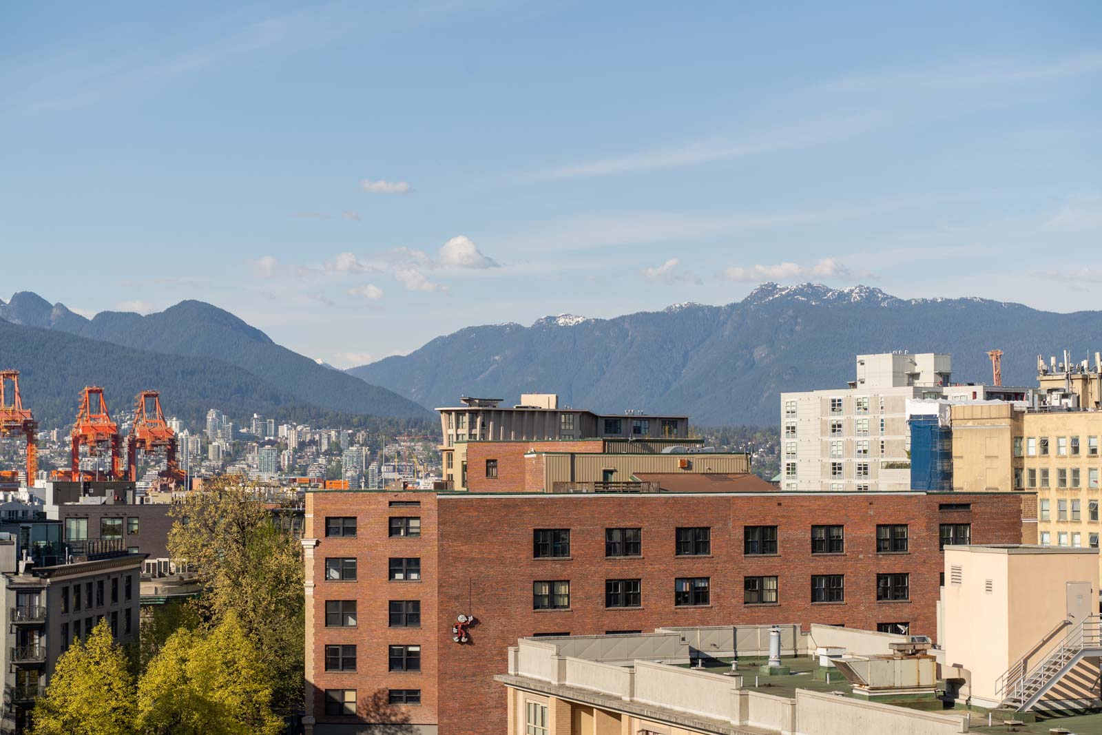Urban cityscape with mid-rise buildings in the foreground, orange cranes to the left, and forested mountains in the background under a blue sky.