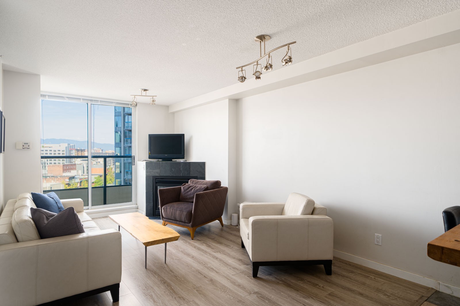 Modern living room with light wood flooring, white walls, cream sofas, an armchair, a fireplace with a TV, large window, and a view of city buildings outside.