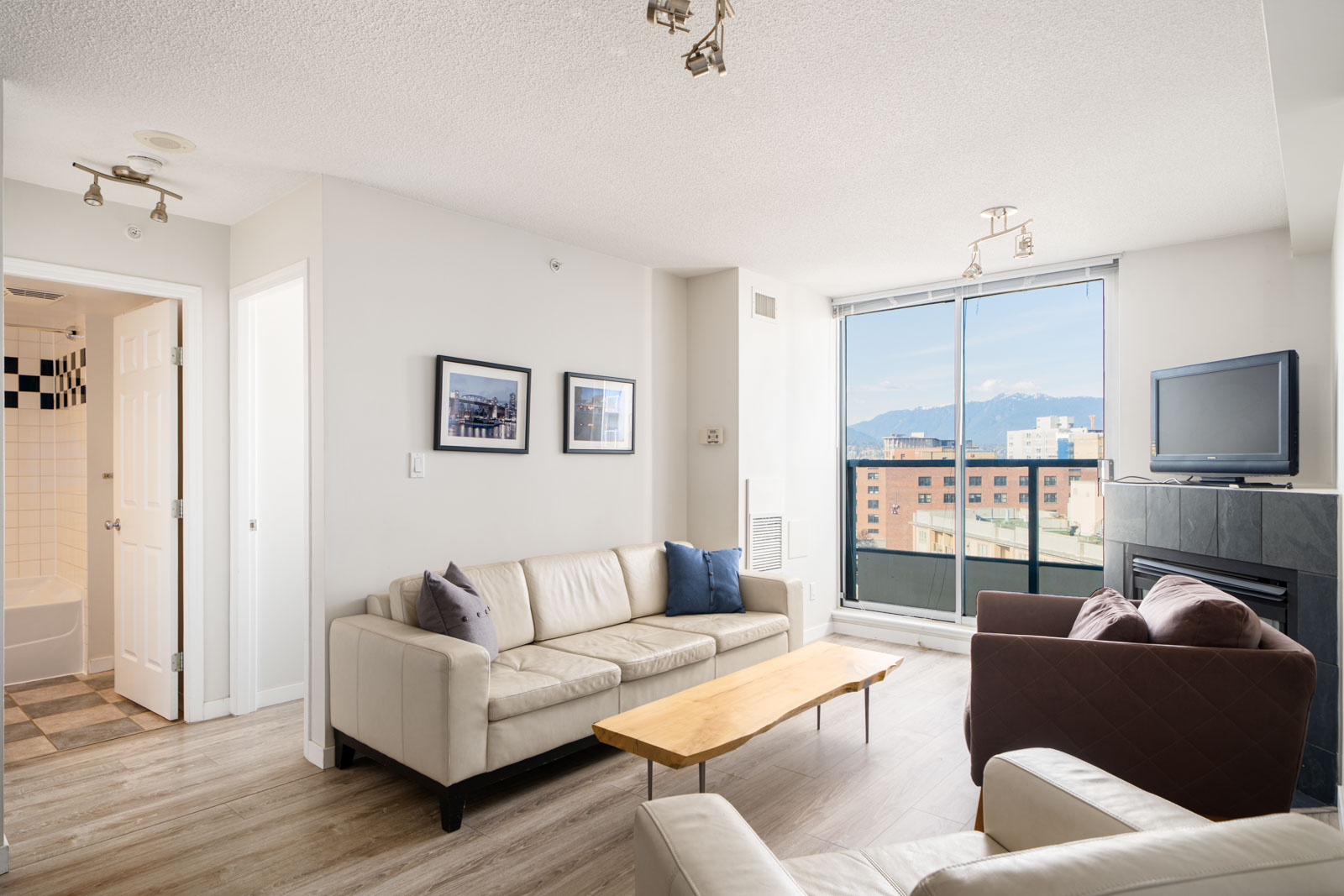 Bright living room with light-colored sofas, a wooden coffee table, wall art, TV above a fireplace, and large windows showing city buildings and mountains in the background.