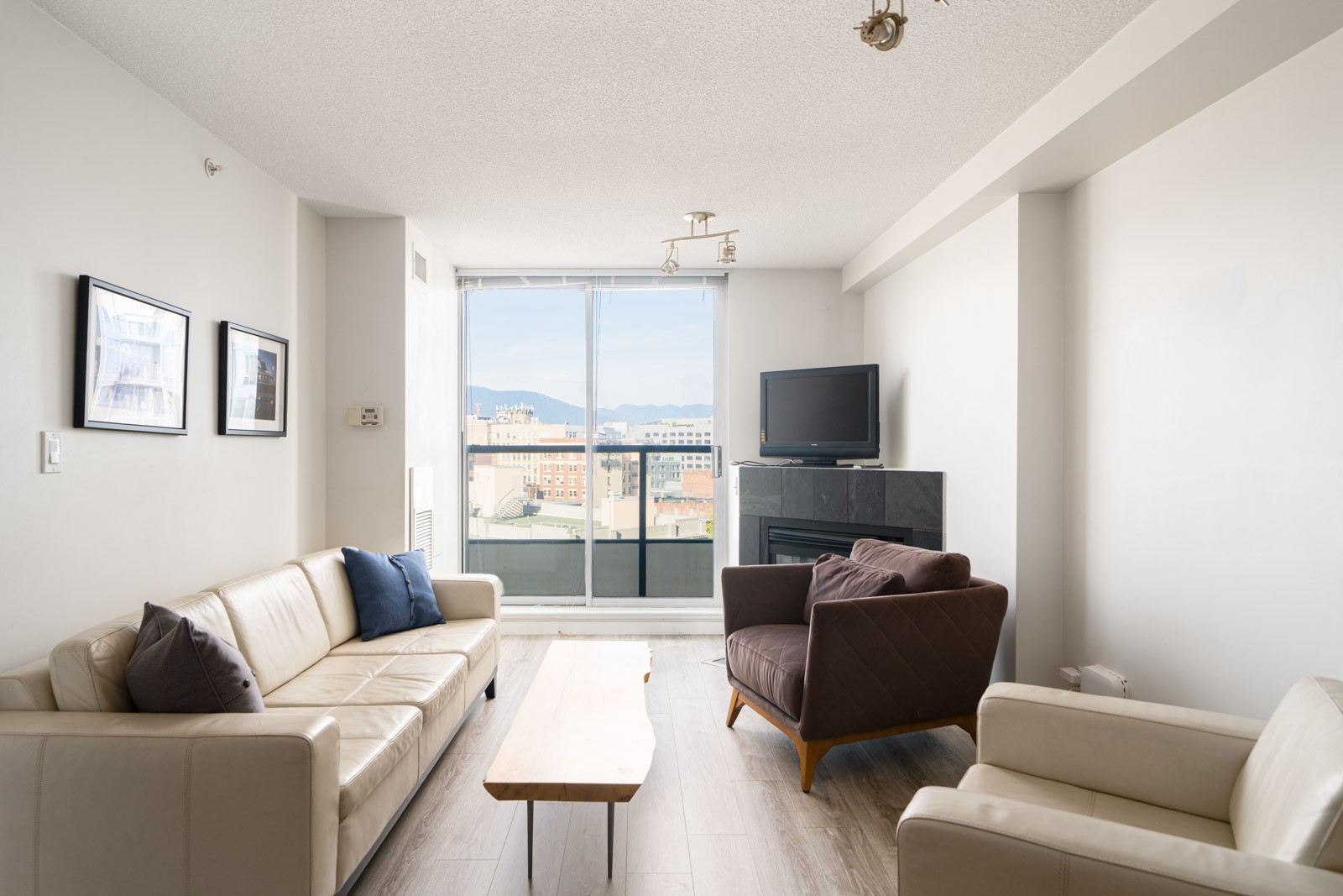 A modern living room with two beige sofas, a brown armchair, a wooden coffee table, a TV on a black stand, large windows, and city views outside.