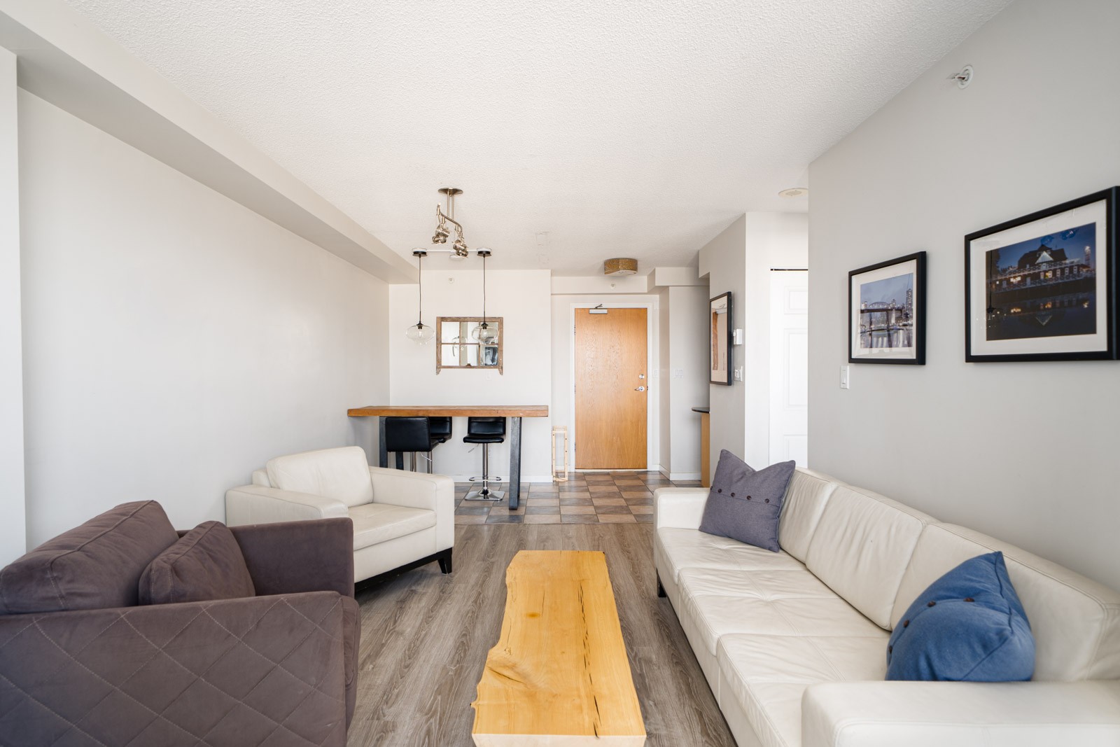 Modern living room with light-colored sofas, a wooden coffee table, wall art, and a view of the entry door and kitchen counter with bar stools in the background.
