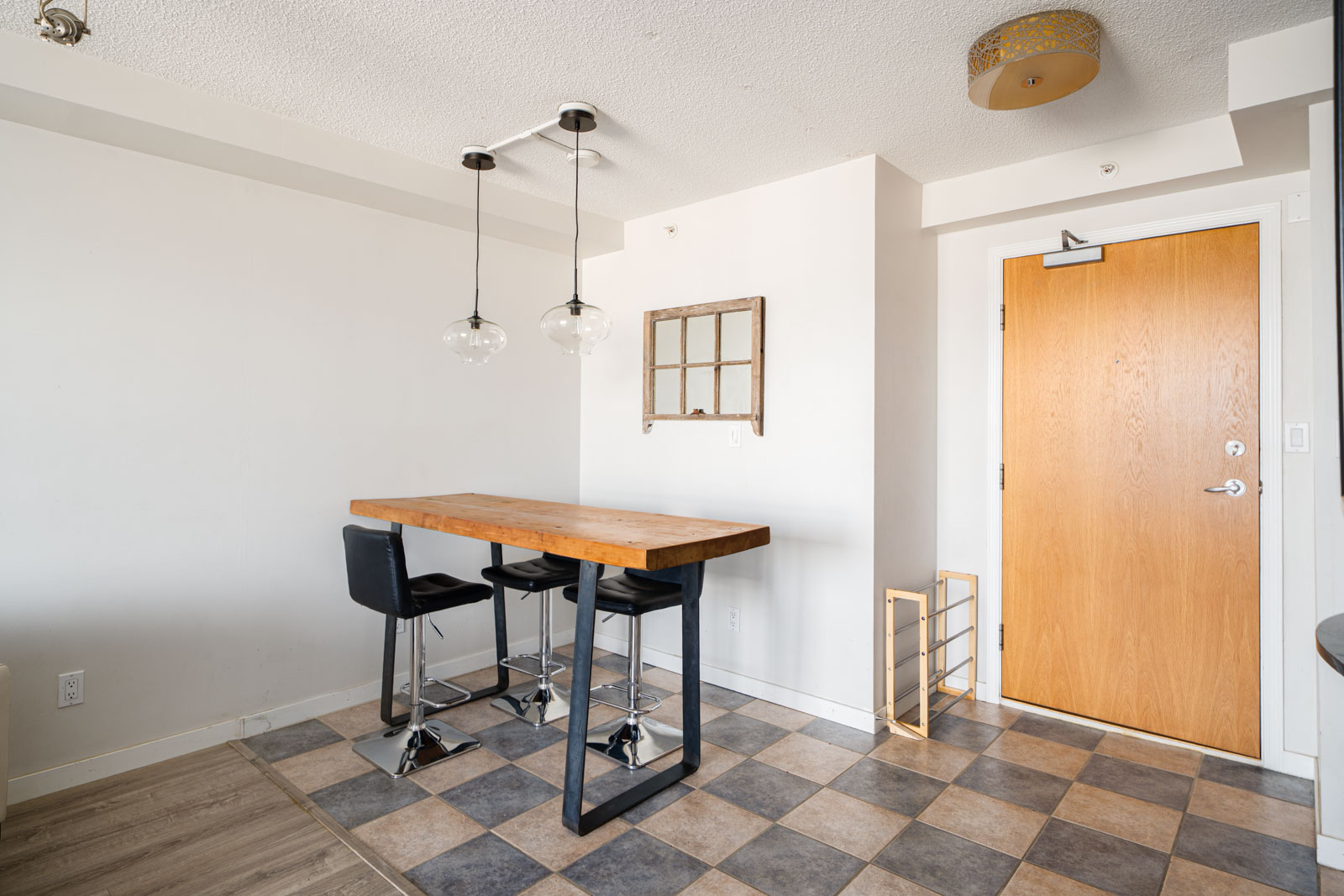 A small dining area with a wooden table, two black chairs, pendant lights, a wall mirror, and a wooden door, on a checkered tile floor.