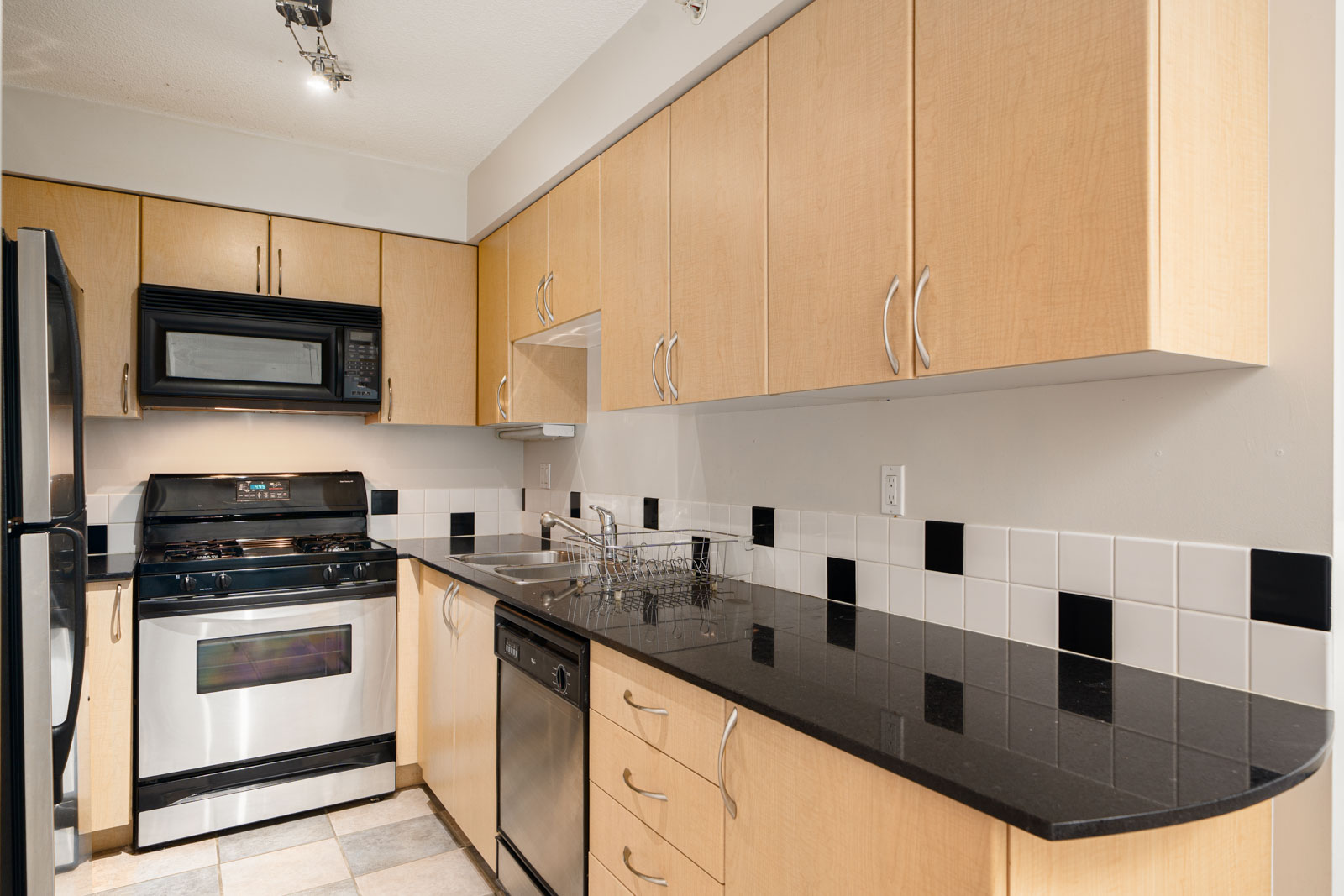 Modern kitchen with light wood cabinets, black countertops, stainless steel appliances, gas stove, dishwasher, and tiled backsplash with black and white accents.