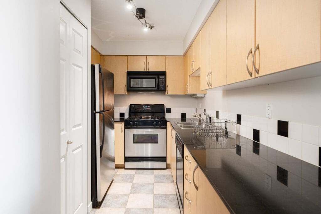 Narrow kitchen with light wood cabinets, black countertops, stainless steel appliances, gas stove, and checkered tile floor.