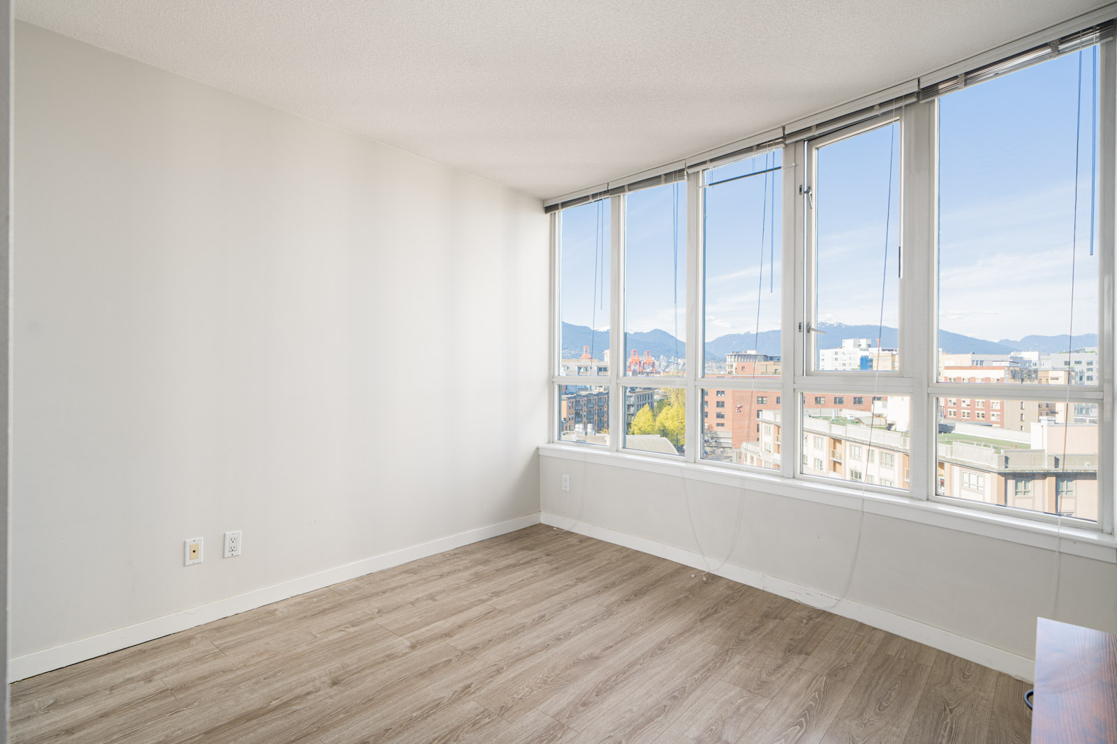 Empty room with light wood flooring, white walls, and large windows offering a city and mountain view.