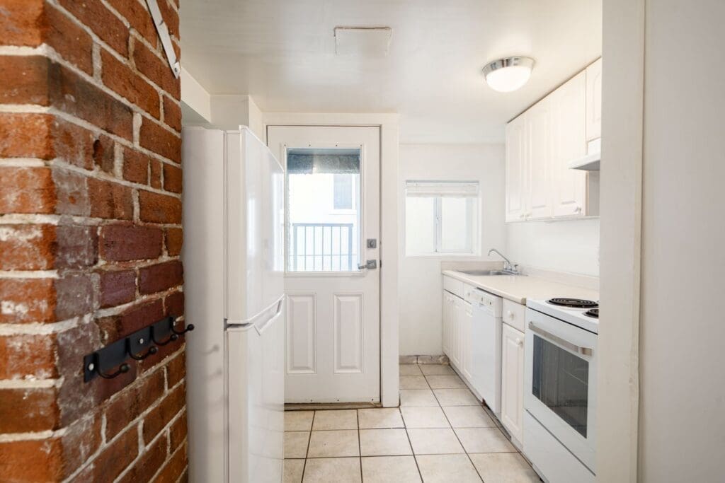 Narrow kitchen with white cabinets, appliances, tiled floor, exposed brick wall, and a door with a window leading outside.
