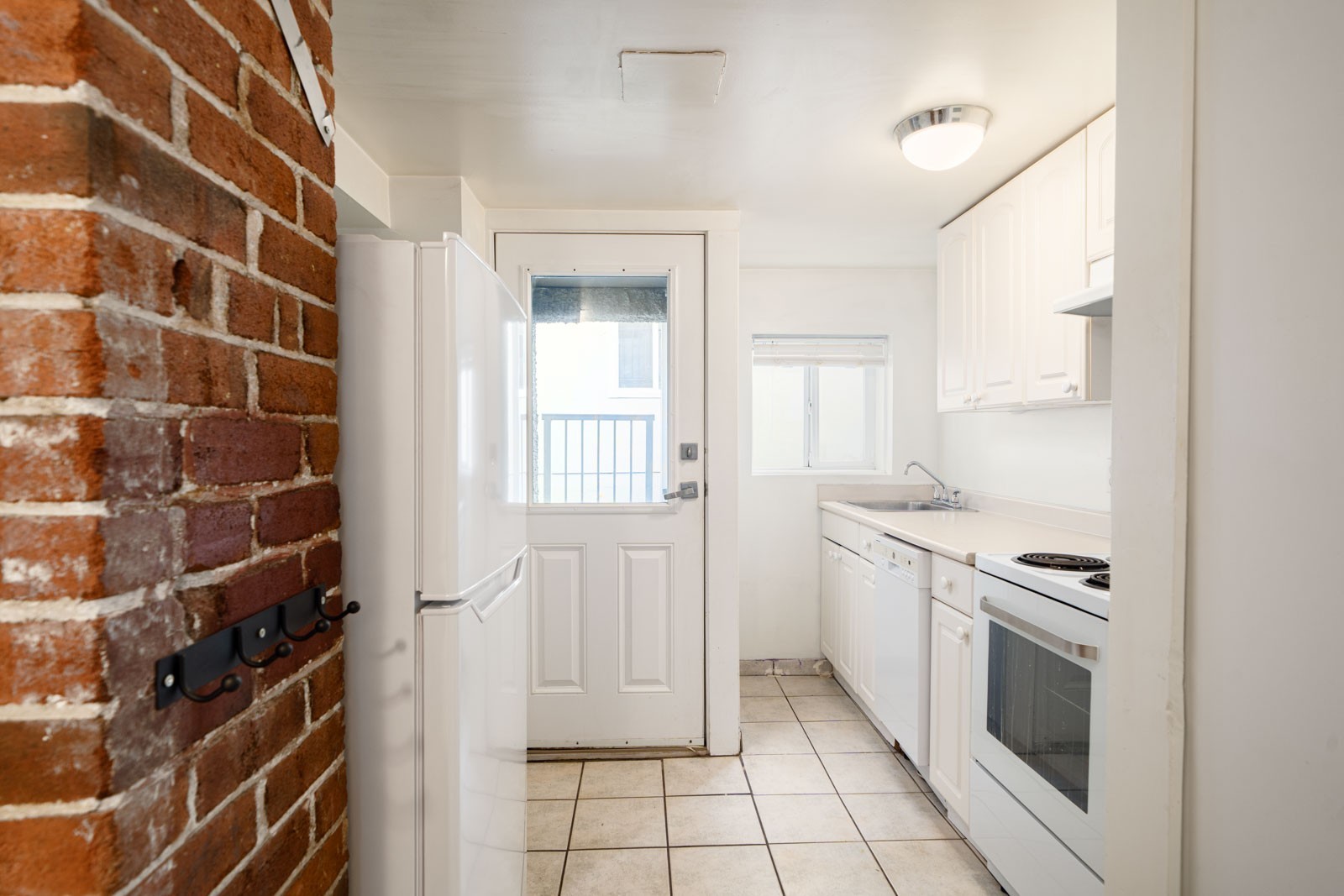Narrow kitchen with white cabinets, appliances, tiled floor, exposed brick wall, and a door with a window leading outside.