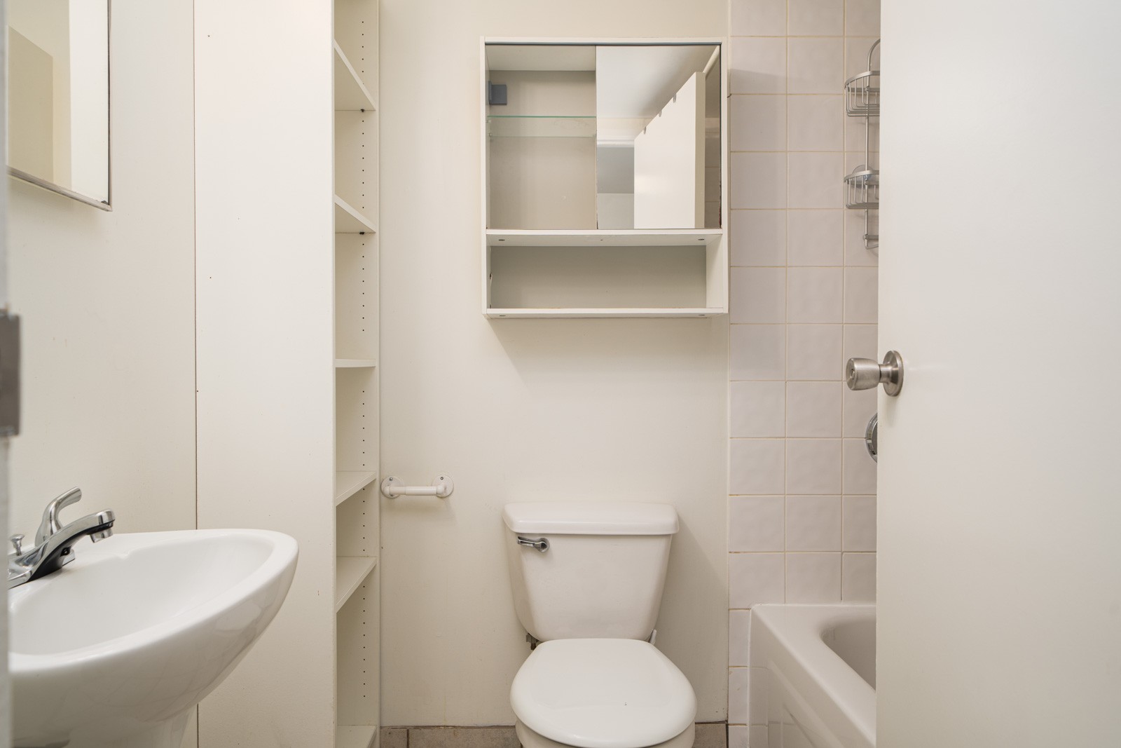 Small bathroom with a white sink, toilet, open shelves, mirrored cabinet, and a bathtub with a tiled wall.