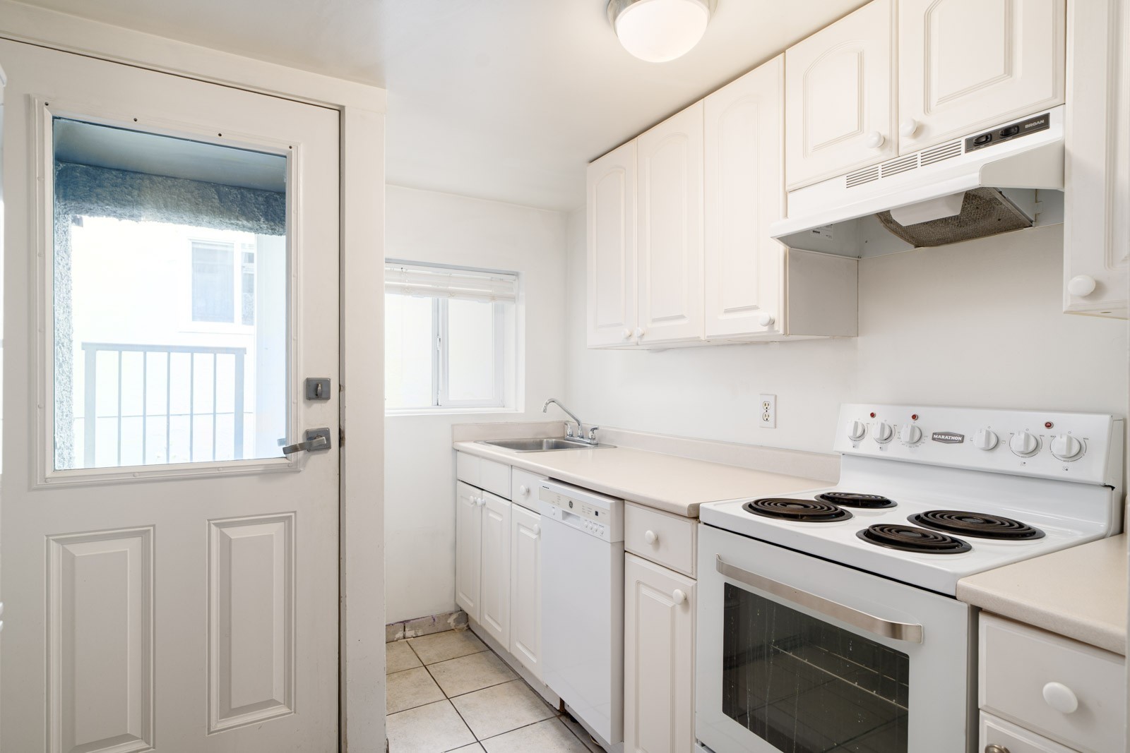 Narrow kitchen with white cabinets, electric stove, dishwasher, small sink, and exterior door. Natural light enters through a window and door.