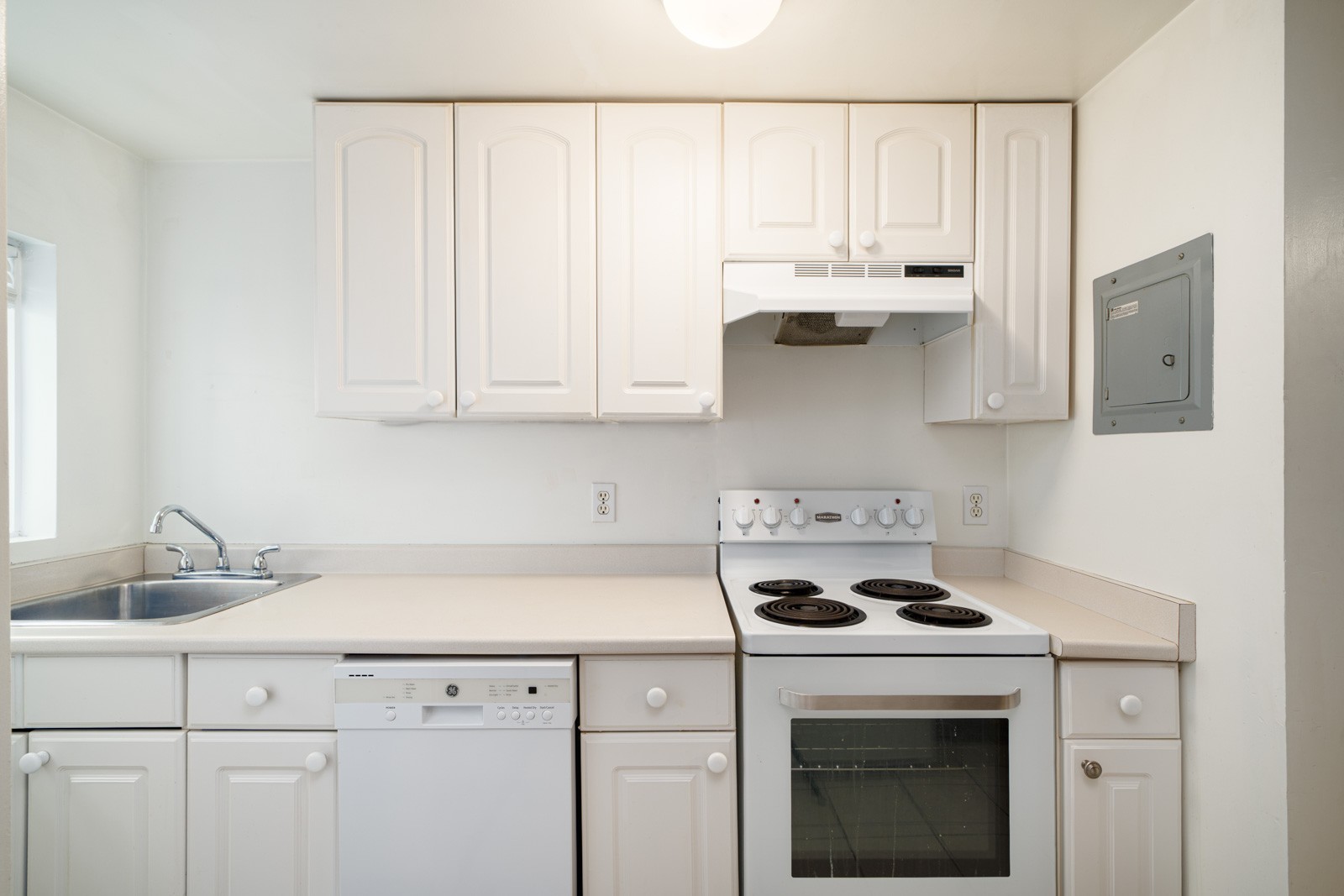 Small kitchen with white cabinets, electric stove, oven, dishwasher, and single sink. Neutral countertops and a gray electrical panel on the wall.