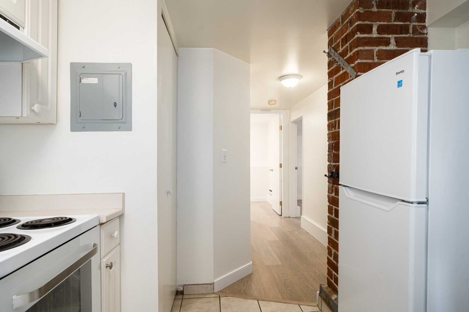 A kitchen with a white stove, countertop, refrigerator, exposed brick wall, and an electrical panel, leading to a hallway with wooden flooring and white walls.