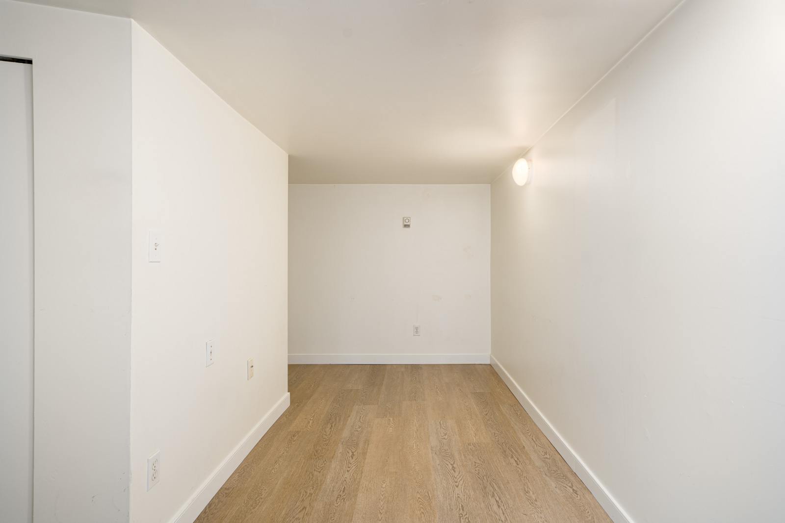 Small, empty room with white walls, light wood flooring, a single light fixture on the right wall, and a partially visible door on the left.