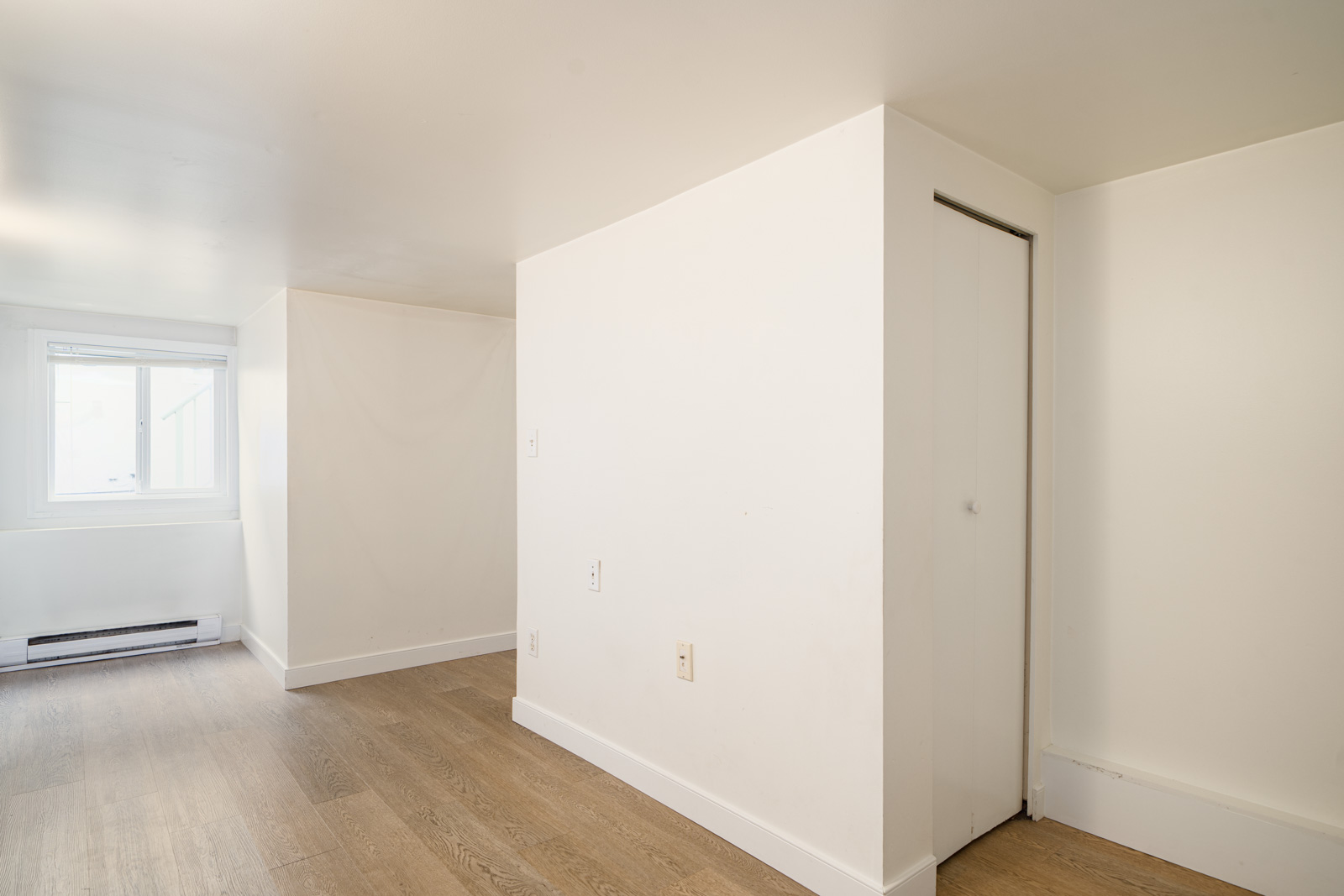 A small, empty room with white walls, light wood flooring, a closed closet door, and a window letting in natural light.