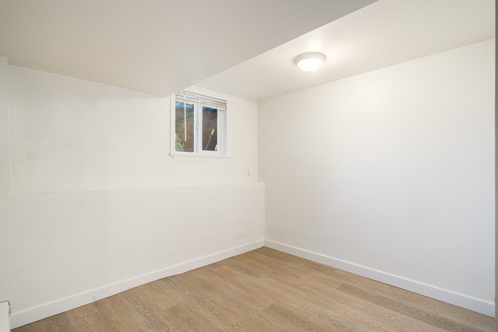 Empty room with white walls, a small window, wood-style flooring, and a ceiling light fixture.