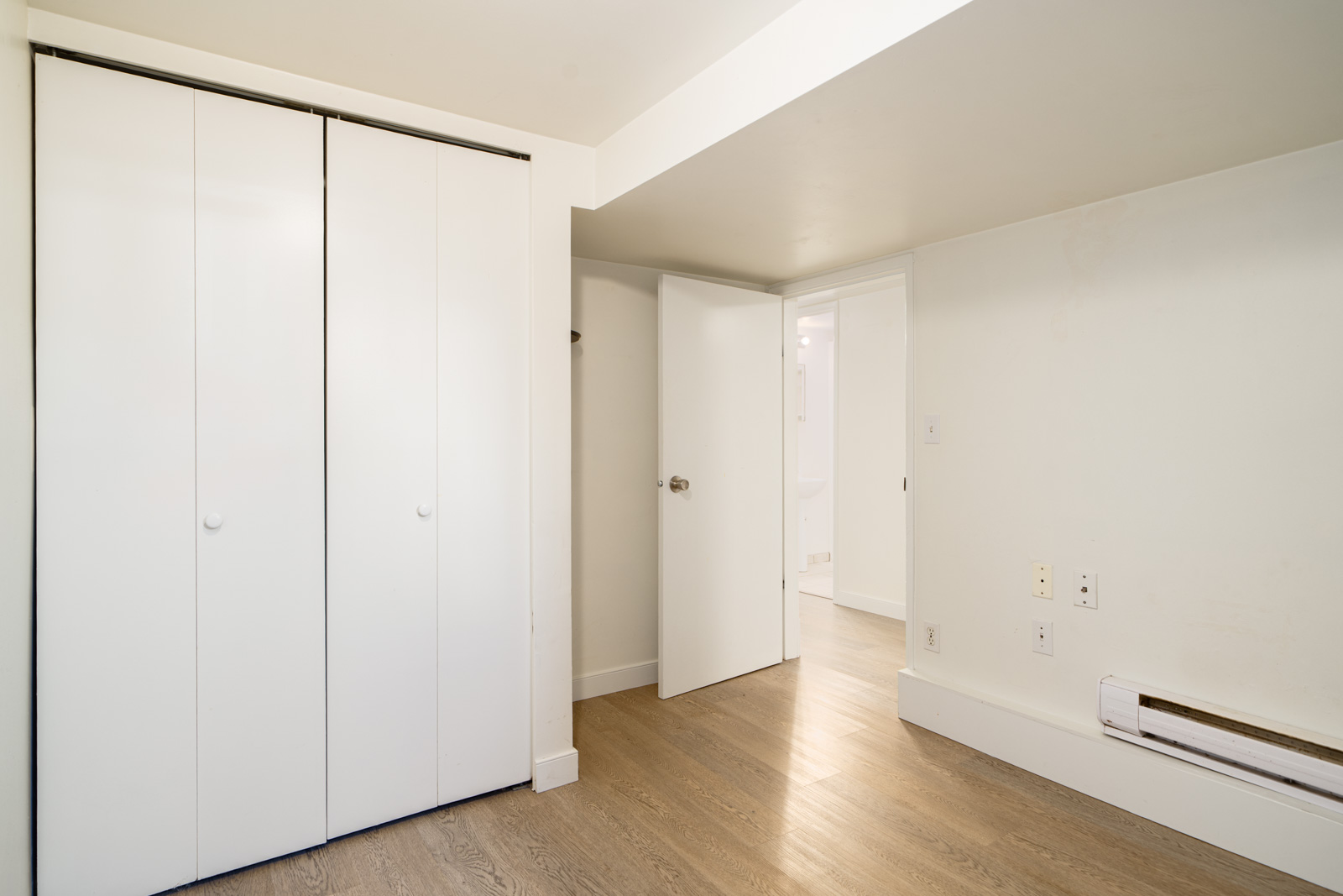 Empty room with light wood flooring, white walls, a closed double-door closet, an open door leading to a bathroom, and a baseboard heater on the right wall.