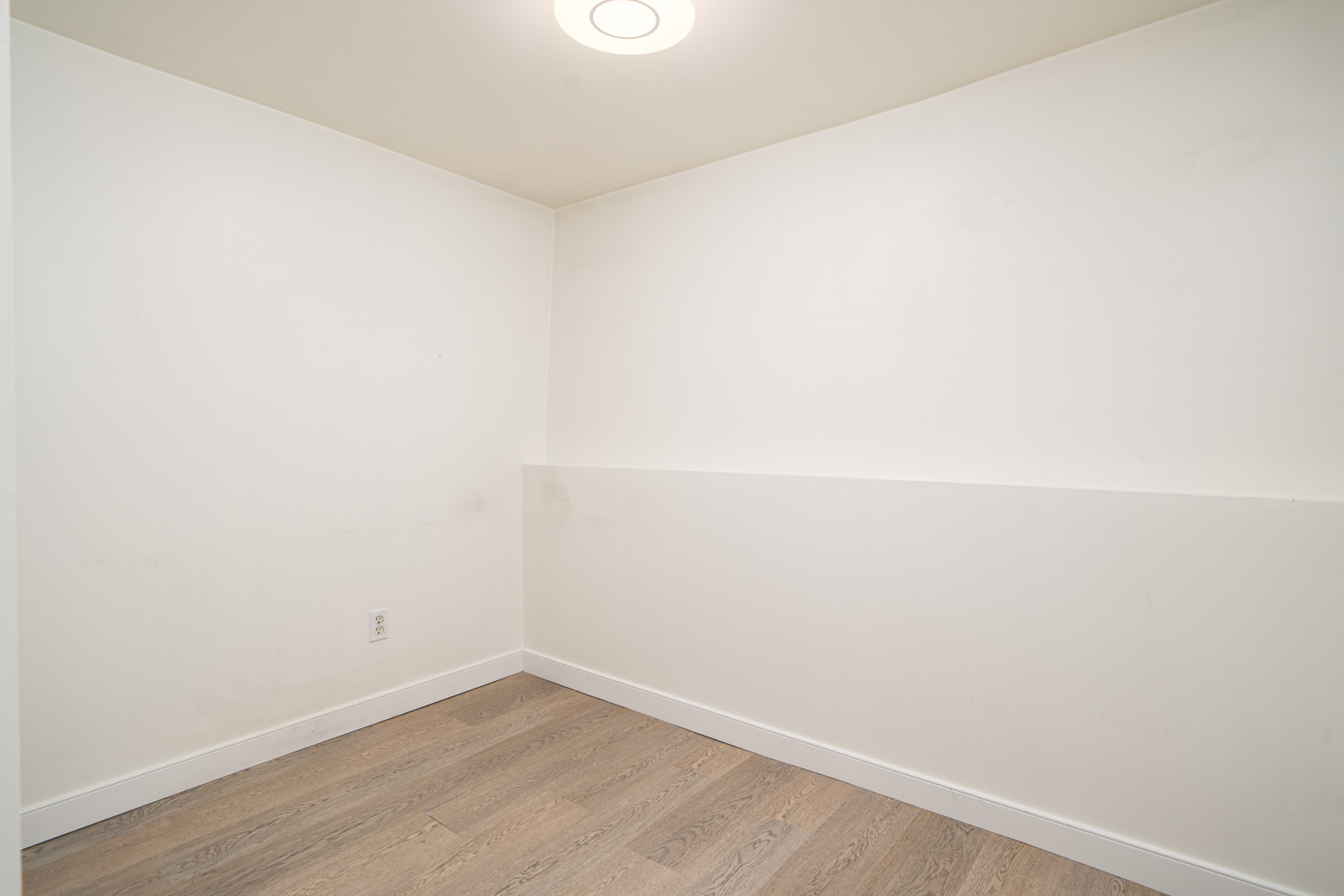 Empty room with white walls, light wood flooring, a single ceiling light, and an electrical outlet on the lower left wall.