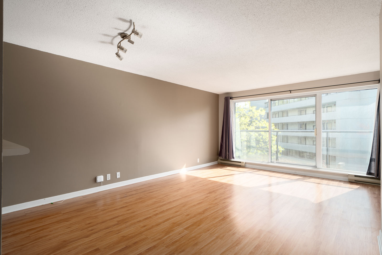 Empty living room with wood flooring, beige walls, ceiling light fixture, large window with curtains, and sunlight streaming in.